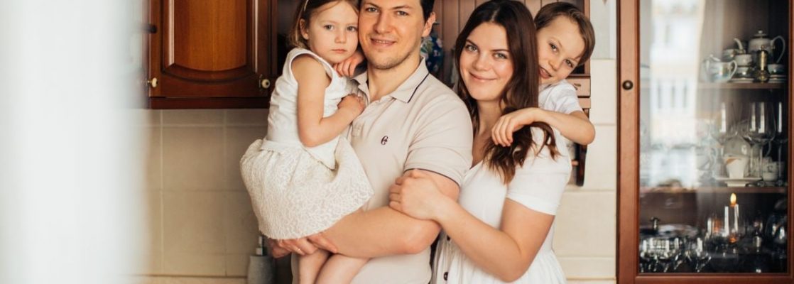 Satisfied man with daughter and woman with son embracing embracing each other while standing against wooden cupboard in kitchen and looking at camera