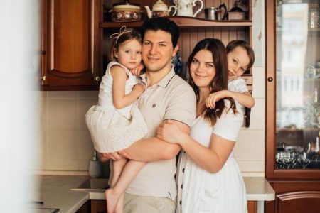Satisfied man with daughter and woman with son embracing embracing each other while standing against wooden cupboard in kitchen and looking at camera
